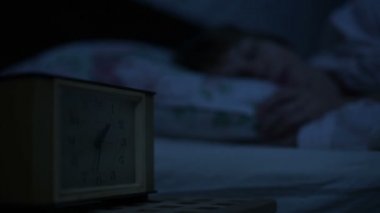 Bir kız yatakta yatarken pijama yakın çekim ve bir alarm saat bakarak uyumak yapamaz. Yüz kızlar için değişken odak saatten. Amerikan gece etkisi. Düşük anahtar soğuk ışık İmitasyon gece