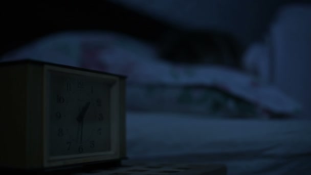 Primer plano de una chica en pijama acostada en la cama e incapaz de dormir mirando un reloj despertador. Enfoque variable de horas a cara con las chicas. El efecto de la noche americana. Noche de imitación de luz fría de llave baja — Vídeos de Stock