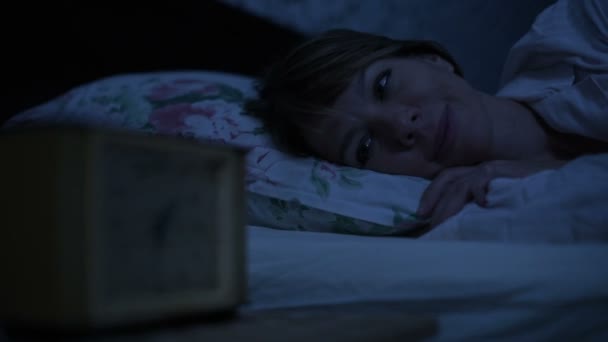 Szczegół dziewczyny w piżamie, leżąc w łóżku i nie może spać, patrząc na budzik. Zmiennej ostrości od godziny do twarzy dziewczyny. Efekt amerykański nocy. Imitacja noc niski klucz światła zimnego — Wideo stockowe