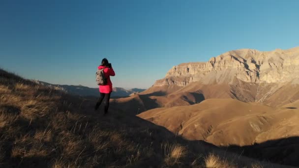 Luftaufnahme eines Reisenden mit Rucksack und Kamera, der bei Sonnenuntergang in den Bergen fotografiert. Fotografin von hinten. die Kamera geht vorbei und fliegt in die Berge — Stockvideo