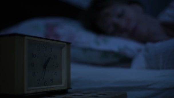 Primer plano de una chica en pijama acostada en la cama e incapaz de dormir mirando un reloj despertador. Enfoque variable de horas a cara con las chicas. El efecto de la noche americana. Noche de imitación de luz fría de llave baja — Vídeos de Stock