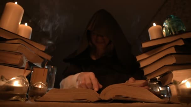 Közepes közeli lány bűvész egy kapucnis, egy sötét szobában, gyertyafény és keres egy varázslat lapozgatott egy könyvet. Alacsony kulcs. Misztikus kis Dof — Stock videók
