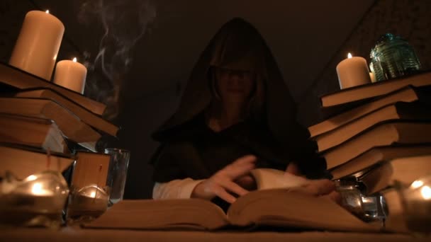 Medium close-up girl magician in a hood in a dark room by candlelight and looking for a spell turning over a book. Low key. Mystic Small DOF — Stock Video