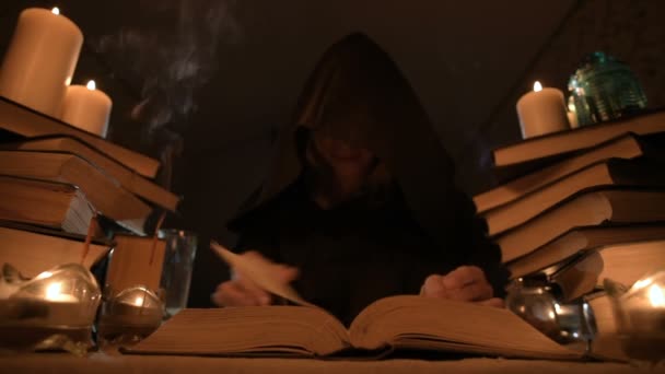 Μέτρια γκρο πλαν κορίτσι μάγος με κουκούλα σε ένα σκοτεινό δωμάτιο από το φως των κεριών και ψάχνει για ένα ξόρκι στροφή πάνω από ένα βιβλίο. Χαμηλών τόνων. Μυστικιστής μικρό Dof — Αρχείο Βίντεο