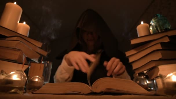 Средний крупный план девочка-фокусница в капюшоне в темной комнате при свечах и ищет заклинание, переворачивающее книгу. Низко. Мистический малый DOF — стоковое видео