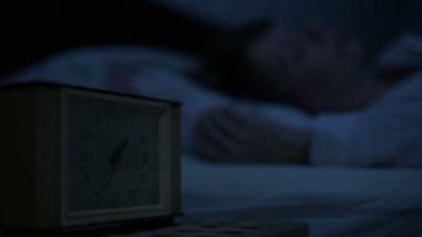 Bir kız yatakta yatarken pijama yakın çekim ve bir alarm saat bakarak uyumak yapamaz. Yüz kızlar için değişken odak saatten. Amerikan gece etkisi. Düşük anahtar soğuk ışık İmitasyon gece