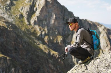 Bir sırt çantası ile güneş gözlüğü sakallı hippi turist mountain yakınındaki dağlarda yüksek bir uçurumun kenarında oturur