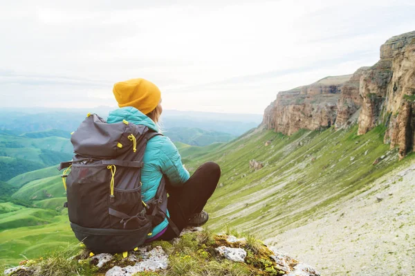 Sentada chica viajero en un sombrero amarillo y un par de gafas de sol sentado al pie de rocas épicas con una mochila al lado y mira hacia otro lado — Foto de Stock