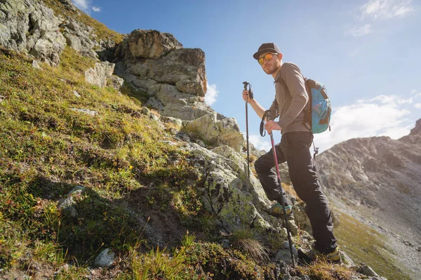 Vousatý muž v sluneční brýle a čepici s batohem stojí na vrcholu skály a dívá do vysoké skalnaté údolí v horách. Koncepce cestovního ruchu a snadný trekking v horách venkovní — Stock fotografie