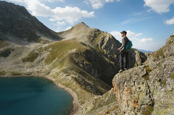 Un turista hipster barbudo en gafas de sol con una mochila está de pie en el borde de un acantilado en las montañas cerca del lago de montaña —  Fotos de Stock