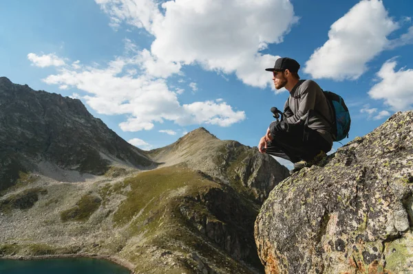 Vousatý bokovky turista v sluneční brýle s batohem na zádech sedí na okraji útesu vysoká v horách poblíž horské jezero — Stock fotografie