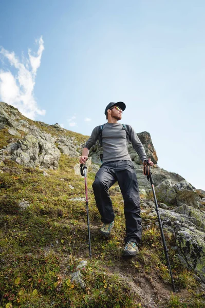 Vousatý muž v sluneční brýle a čepici s batohem stojí na vrcholu skály a dívá do vysoké skalnaté údolí v horách. Koncepce cestovního ruchu a snadný trekking v horách venkovní — Stock fotografie