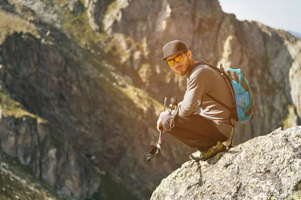 Vousatý bokovky turista v sluneční brýle s batohem na zádech se nachází na okraji útesu vysoká v horách poblíž hory — Stock fotografie
