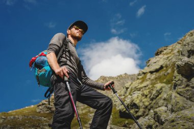 Porter sakal ve bir sırt çantası içinde güneş gözlüğü ile bir şık hipster seyahat ve trekking Polonyalılar bir rock Dağları'nda yamaç ve mavi gökyüzü arka planı üzerinde duran bir kap