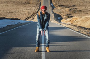 Güneş gözlüğü vintage bir sırt çantası ile şık bir sakallı hippi güneşli bir günde bir ülke yol asfalt üzerinde duruyor. Otostop ve hiking kavramı