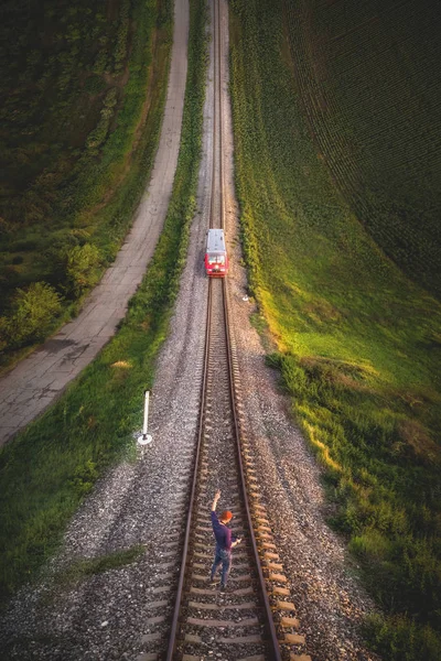 Aerial view A male hipster in a cap holding up a hand with a gesture Victory is standing on rails in front of an approaching train. Reverse vertical panorama in the style of Inception — Stock Photo, Image