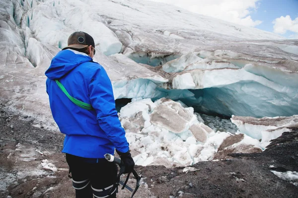 Молодой проводник в кепке с альпинистским снаряжением и ледорубом в руке стоит на фоне рассыпающегося ледника и трещин. Концепция глобального потепления и профессиональные гиды — стоковое фото
