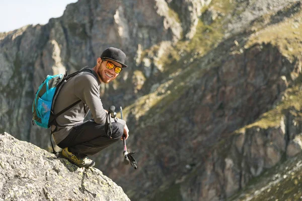 Vousatý bokovky turista v sluneční brýle s batohem na zádech se nachází na okraji útesu vysoká v horách poblíž hory — Stock fotografie