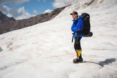 Gezgin bir kap ve gökyüzü ve bulutlar karşı buzul üzerinde karlı dağlarda onun omuzlarında bir sırt çantası ile güneş gözlüğü. Gezgin bir doğal ortamda
