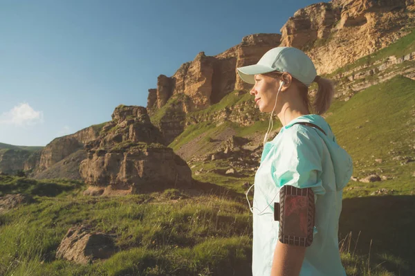 Attractive sports girl in a cap and headphone before jogging in a picturesque location next to the rocks at sunset. Bekerja di luar ruangan. Tampilan belakang — Stok Foto