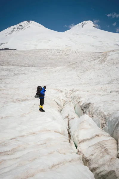 Viaggiatore in berretto e occhiali da sole con uno zaino sulle spalle tra le montagne innevate sul ghiacciaio contro il cielo e le nuvole. Viaggiatore in un ambiente naturale — Foto Stock