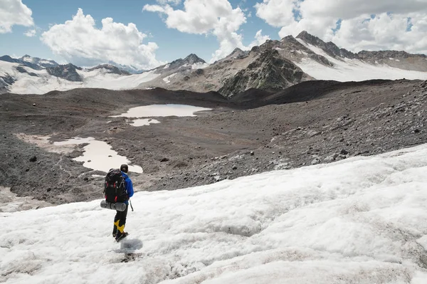 Viajante de boné e óculos de sol com uma mochila nos ombros nas montanhas nevadas no glaciar contra o céu e as nuvens. Viajante em um ambiente natural — Fotografia de Stock