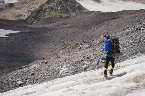 Reiziger in een pet en zonnebril met een rugzak op zijn schouders in de besneeuwde bergen op de gletsjer tegen de lucht en de wolken. Reiziger in een natuurlijke omgeving — Stockfoto
