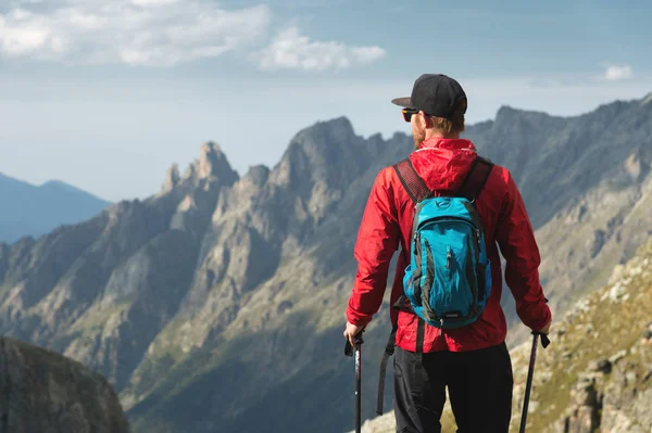 Vousatý muž v sluneční brýle a čepici s batohem stojí na vrcholu skály a dívá do vysoké skalnaté údolí v horách. Koncepce cestovního ruchu a snadný trekking v horách venkovní — Stock fotografie