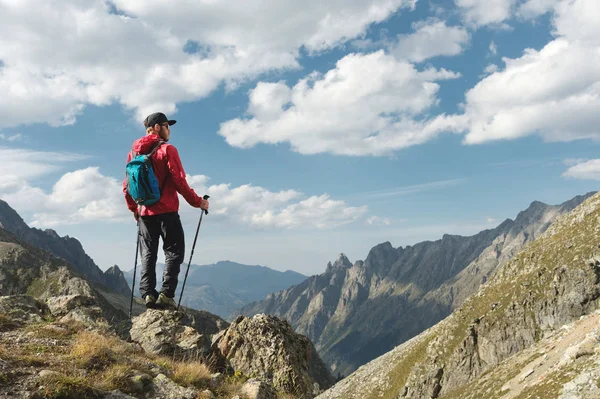 Vousatý muž v sluneční brýle a čepici s batohem stojí na vrcholu skály a dívá do vysoké skalnaté údolí v horách. Koncepce cestovního ruchu a snadný trekking v horách venkovní — Stock fotografie