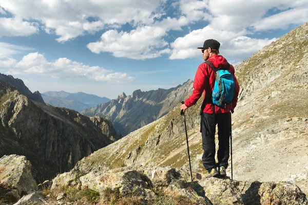 Vousatý muž v sluneční brýle a čepici s batohem stojí na vrcholu skály a dívá do vysoké skalnaté údolí v horách. Koncepce cestovního ruchu a snadný trekking v horách venkovní — Stock fotografie