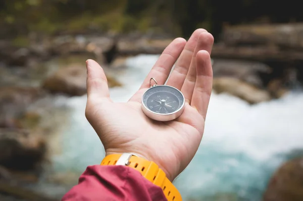 Uma bela mão masculina com uma pulseira de relógio amarela segura uma bússola magnética em uma floresta de outono conífera contra um rio de montanha com pedras rochosas. O conceito de encontrar o caminho e a verdade — Fotografia de Stock