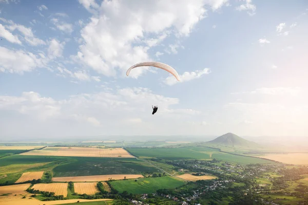 Professional paraglider in a cocoon suit flies high above the ground against the sky and fields with mountains — Stock Photo, Image