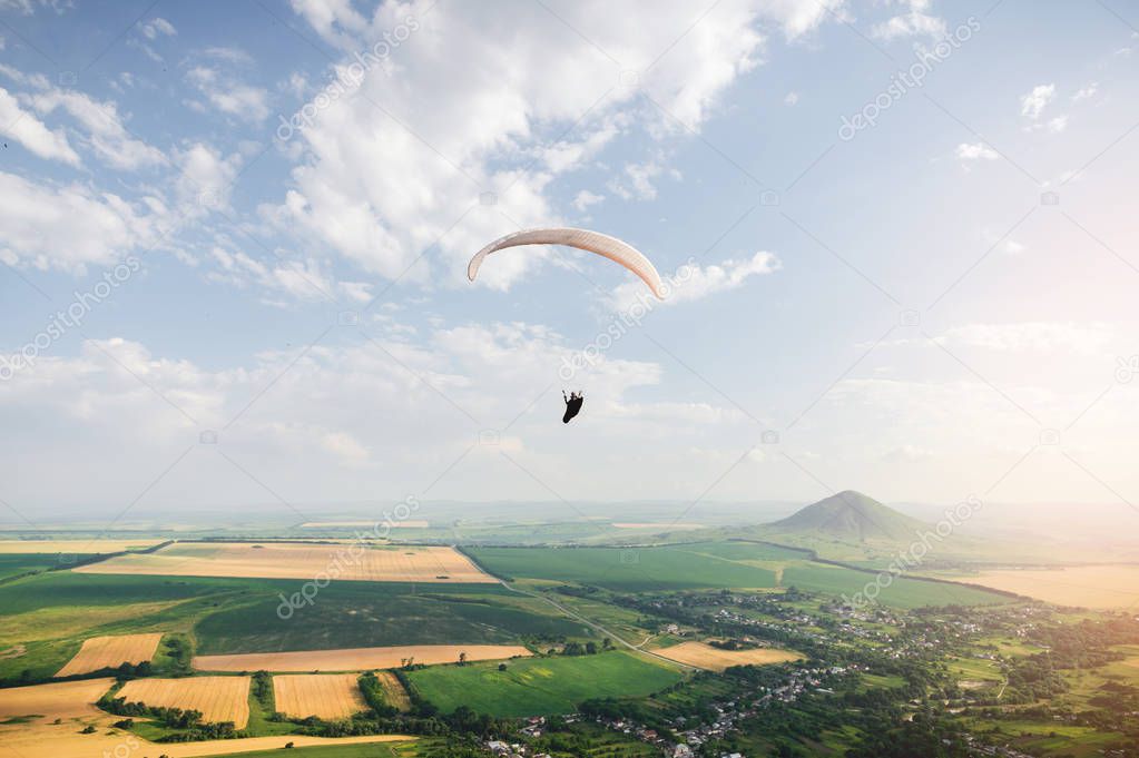 Professional paraglider in a cocoon suit flies high above the ground against the sky and fields with mountains