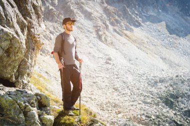 Porter sakal ve bir sırt çantası içinde güneş gözlüğü ile bir şık hipster seyahat ve bir kap trekking Polonyalılar ile bir kayaya bir dağ sirk zemin üzerinde duruyor