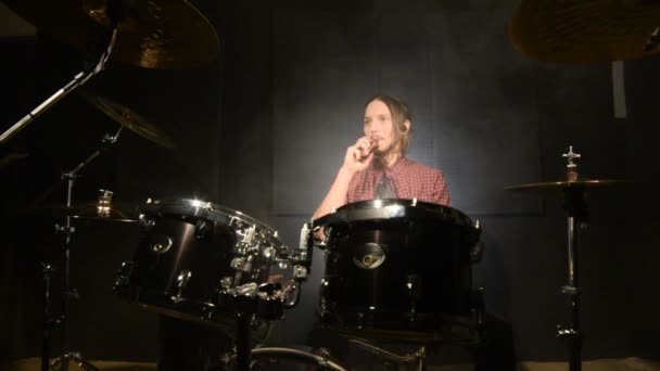 A long-haired drummer sitting behind a drum kit is hovering with a vape. Vape culture in music. Static plan — Stock Video