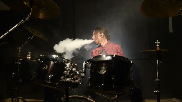 Een langharige drummer zitten achter een drumstel is met een vape zweven. Vape cultuur in muziek. Statische plan — Stockvideo