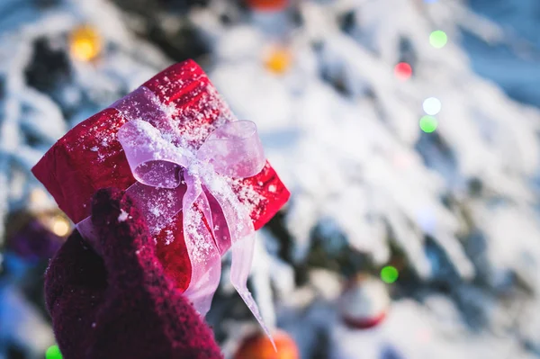 Close-up. Female hand holding red Christmas gift in red mittens in the forest on the background of the New Year tree. The concept of receiving Christmas gifts — Stock Photo, Image