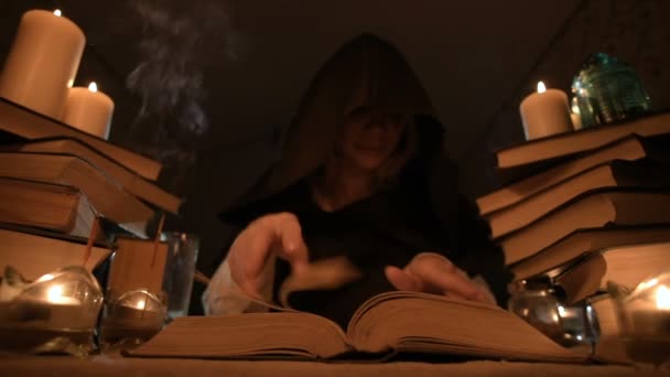 Middellange close-up meisje tovenaar in een kap in een donkere kamer door kaarslicht en op zoek naar een spreuk draaien meer dan een boek. Lage sleutel. Mystic kleine Dof — Stockvideo