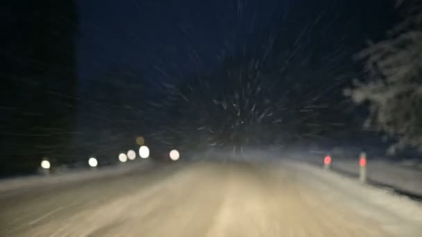 私たちは夜に雪に覆われた道の冬の森をドライブします。フロント ガラスに飛んで雪の結晶に焦点を当てる — ストック動画