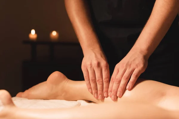 Close-up van mannelijke handen doen kalf massage van vrouwelijke benen in een donkere kamer met kaarsen op de achtergrond. Cosmetologie en wellness-behandelingen — Stockfoto