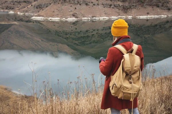 Un ritratto dal retro di una ragazza fotografo viaggiatore sullo sfondo di un lago in montagna in autunno o all'inizio della primavera. Concetto di viaggio — Foto Stock