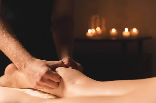 Close-up de mãos masculinas fazendo massagem na panturrilha de pernas femininas em um quarto escuro com velas no fundo. Cosmetologia e tratamentos de spa — Fotografia de Stock