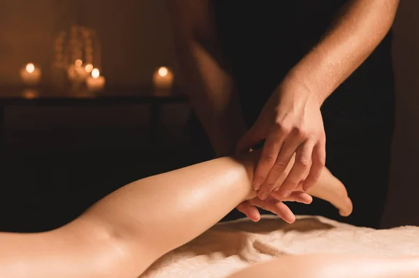 Primer plano de las manos masculinas haciendo masaje de pantorrilla de piernas femeninas en una habitación oscura con velas en el fondo. Cosmetología y tratamientos de spa — Foto de Stock