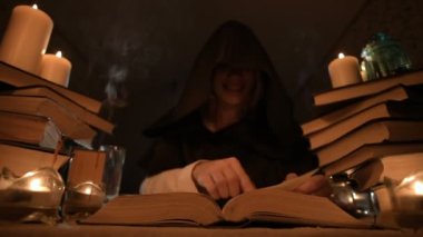 Mum ışığı ve seyir için bir kitap üzerinde bir büyü tarafından karanlık bir odada bir başlıklı orta yakın çekim kız sihirbaz. Düşük anahtar. Mistik küçük Dof