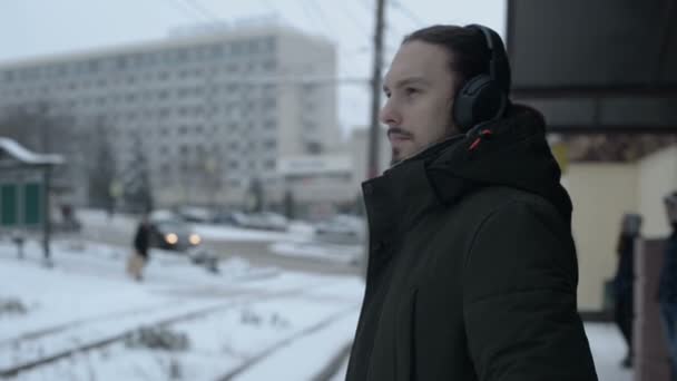 Portrait of a young long-haired man with a beard in headphones standing at a tram stop in winter and waiting for a tram listening to music — Stock Video