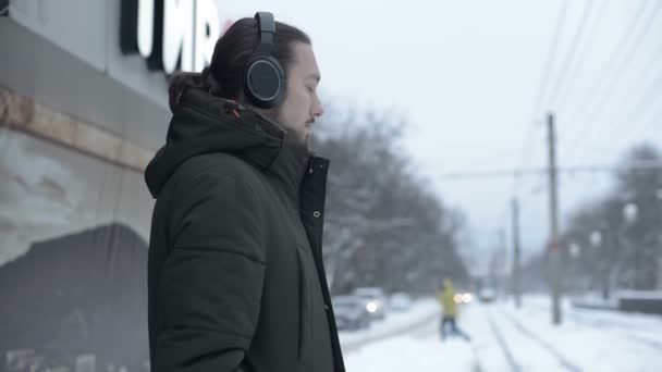 冬に路面電車の停留所で立っていると、音楽を聞いて電車を待ってのヘッドフォンでひげと長髪青年の肖像画 — ストック動画