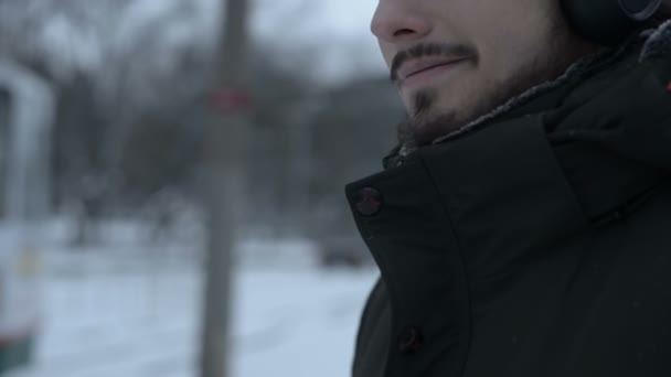 Close-up Retrato de um jovem de cabelos compridos com barba em fones de ouvido em pé em uma parada de bonde no inverno e esperando por um bonde ouvindo música — Vídeo de Stock