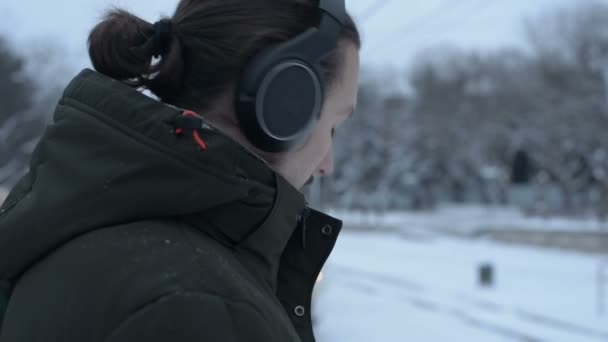 Close-up Retrato de um jovem de cabelos compridos com barba em fones de ouvido em pé em uma parada de bonde no inverno e esperando por um bonde ouvindo música — Vídeo de Stock