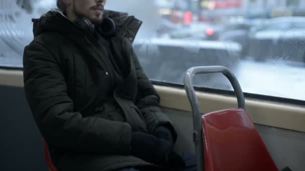 Close-up van portret jonge langharige bebaarde man in een jasje en grote hoofdtelefoon zit in openbaar vervoer door de tram bij het raam en luistert naar muziek of een audioboek in de winter — Stockvideo