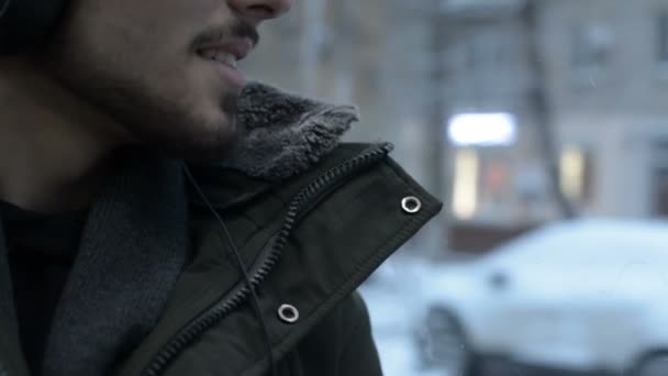 Retrato de cerca Joven hombre barbudo de pelo largo con chaqueta y auriculares grandes se sienta en el transporte público en el tranvía por la ventana y escucha música o un audiolibro en el invierno — Vídeos de Stock